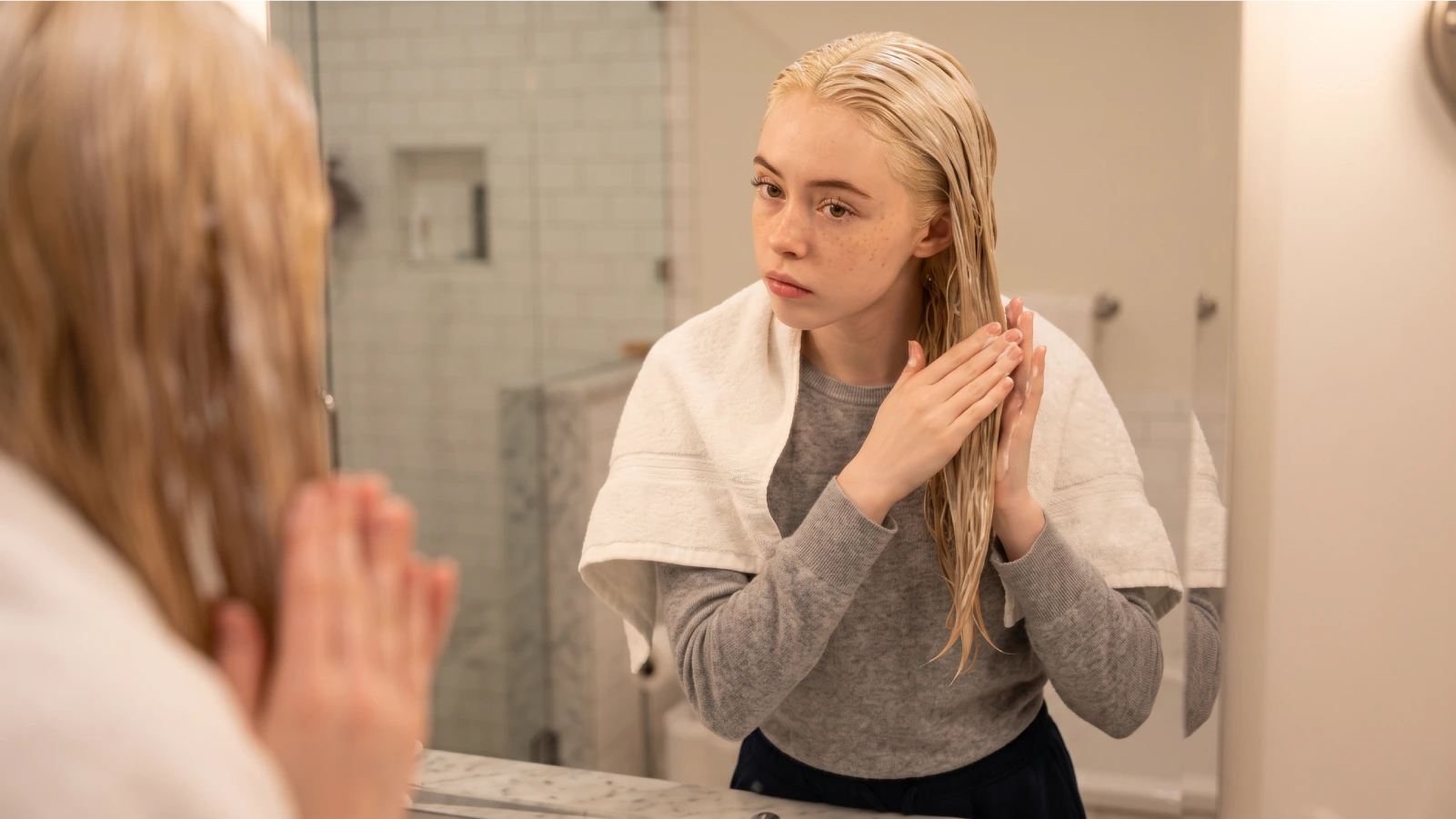 Young woman standing at a mirror applying a conditioning bond treatment through her wet bleached hair, with a towel draped over her shoulders