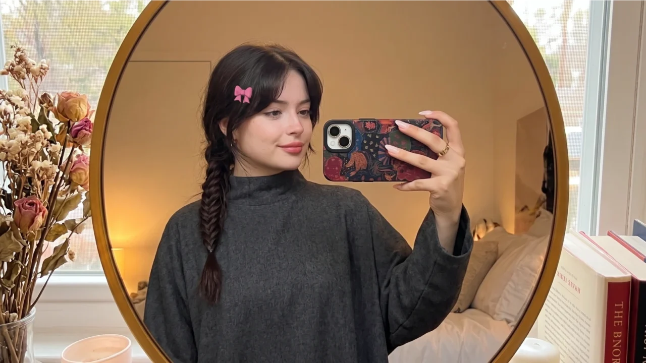 Young woman taking a mirror selfie showing a fishtail braid with a pink bow clip, wearing a cozy oversized sweater in a warm aesthetic bedroom setup