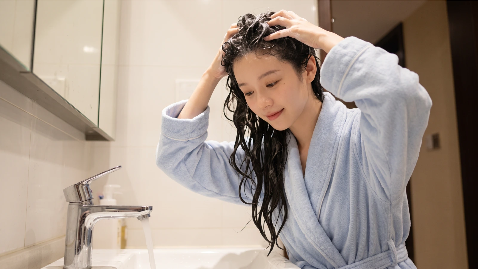 A young woman massaging shampoo into her wet scalp at a bathroom sink, following a proper wavy hair washing routine