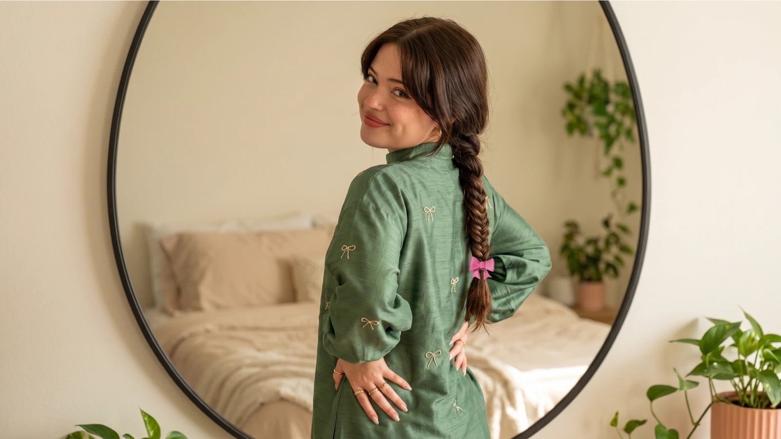 Young woman demonstrating a finished fishtail braid draped over her shoulder, standing in front of a round mirror in a cozy green plant-filled bedroom