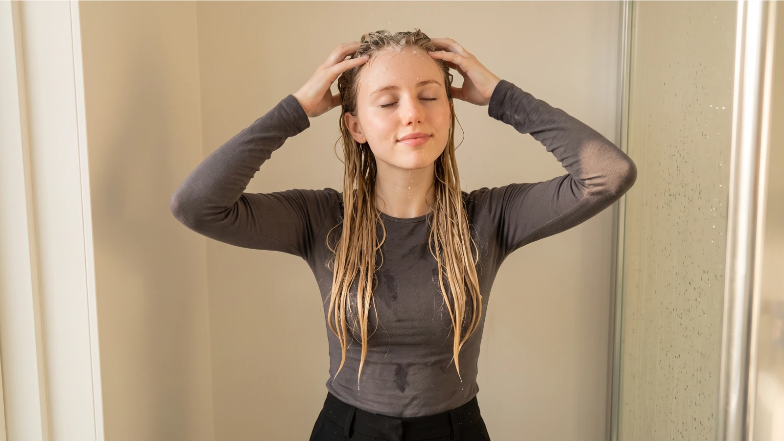 A young woman massaging shampoo into her scalp with both hands while washing her golden blonde colored hair, demonstrating the correct technique to protect hair color during washing